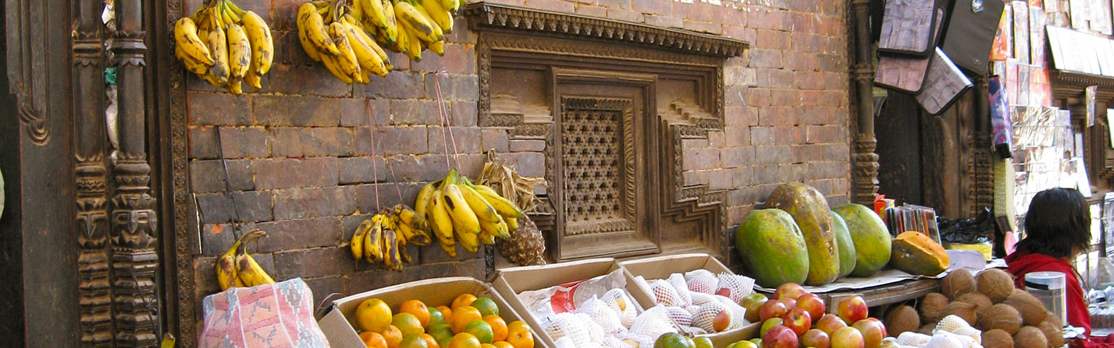 Fruit, including bananas, apples, and mangos, are stacked in boxes and displayed on a table in front of a brick wall. 